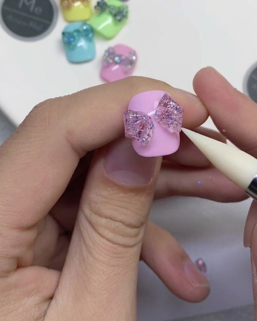 Nahaufnahme eines weißen Silikonwerkzeugs, das in den mittleren Streifen des Glitzergels drückt, um zwei Schlaufen auf einem rosa Nagel zu verbinden.