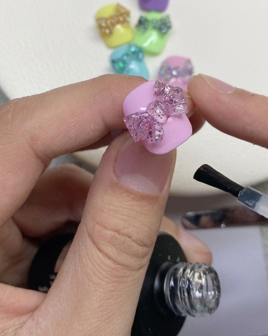 Nahaufnahme eines Nagelpinsels, der einen klaren Überlack um eine 3D-Glitzerschleife auf einem rosa lackierten Nagel aufträgt.