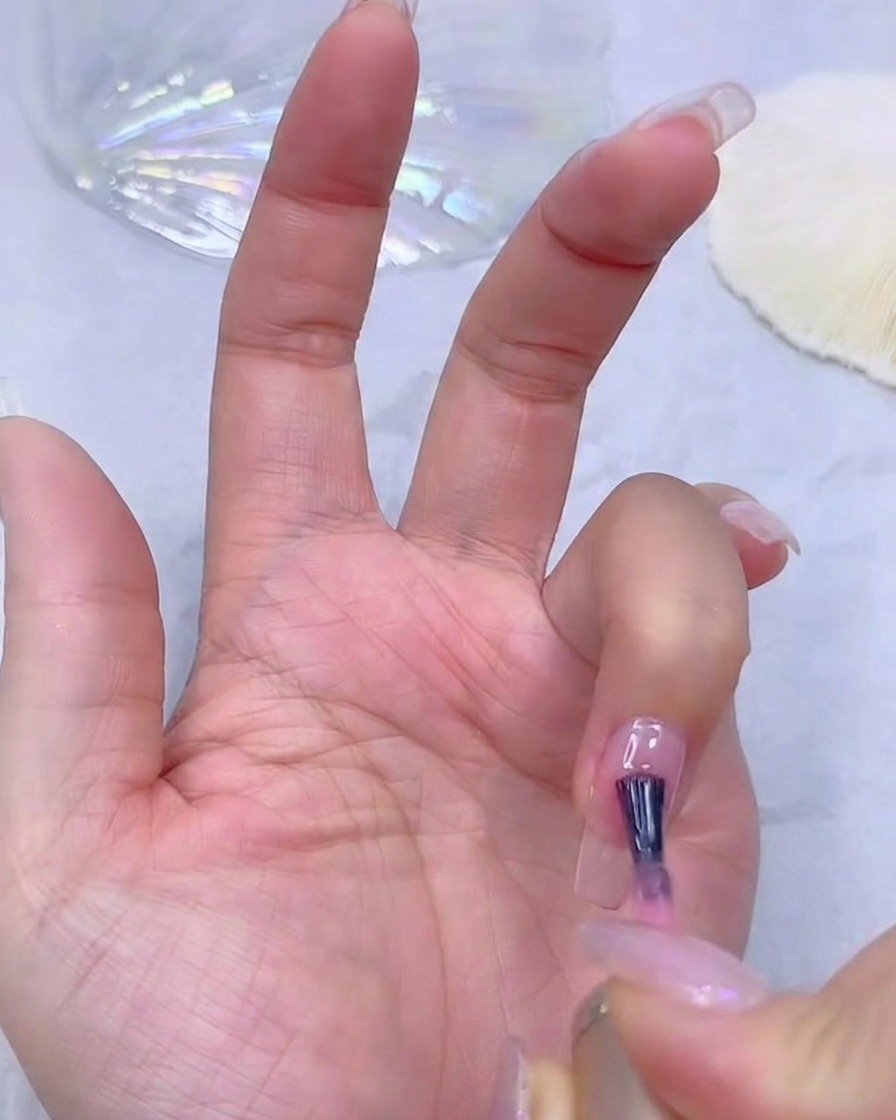 Die Hand einer Nageldesignerin trägt einen transparenten rosa Gel-Base-Coat auf eine klare Nagelverlängerung auf.
