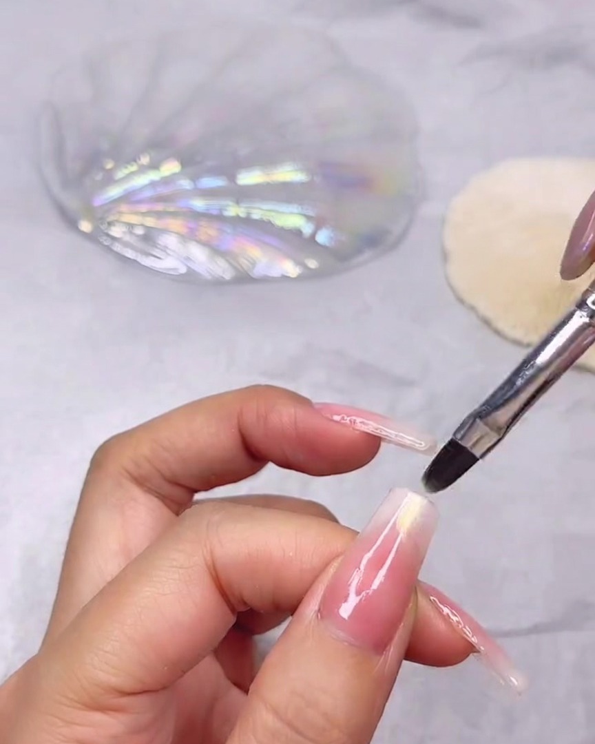 Ein feiner Nagelpinsel trägt schimmerndes, klares Muschelgel auf die Spitze eines langen Nagels mit rosa-weißem Farbverlauf auf.