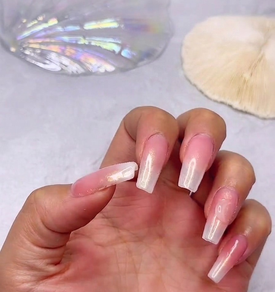 Nahaufnahme einer Hand mit langen, glänzenden Nägeln und einem rosa-weißen Farbverlauf, bei der ein großer, klarer Herz-Strassstein nahe der Nagelhaut platziert ist.