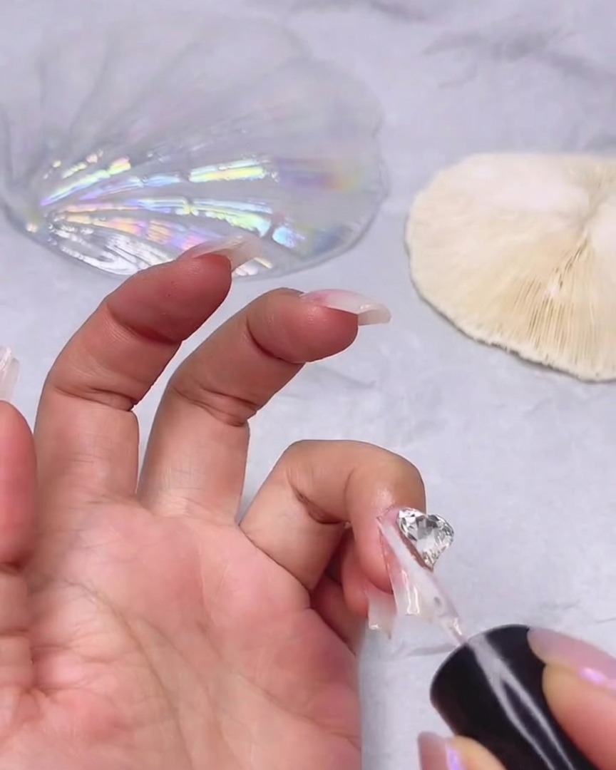 Ein feiner Pinsel trägt ein klares Versiegelungsgel um die Kanten eines großen, herzförmigen, klaren Strasssteins auf dem Fingernagel auf.