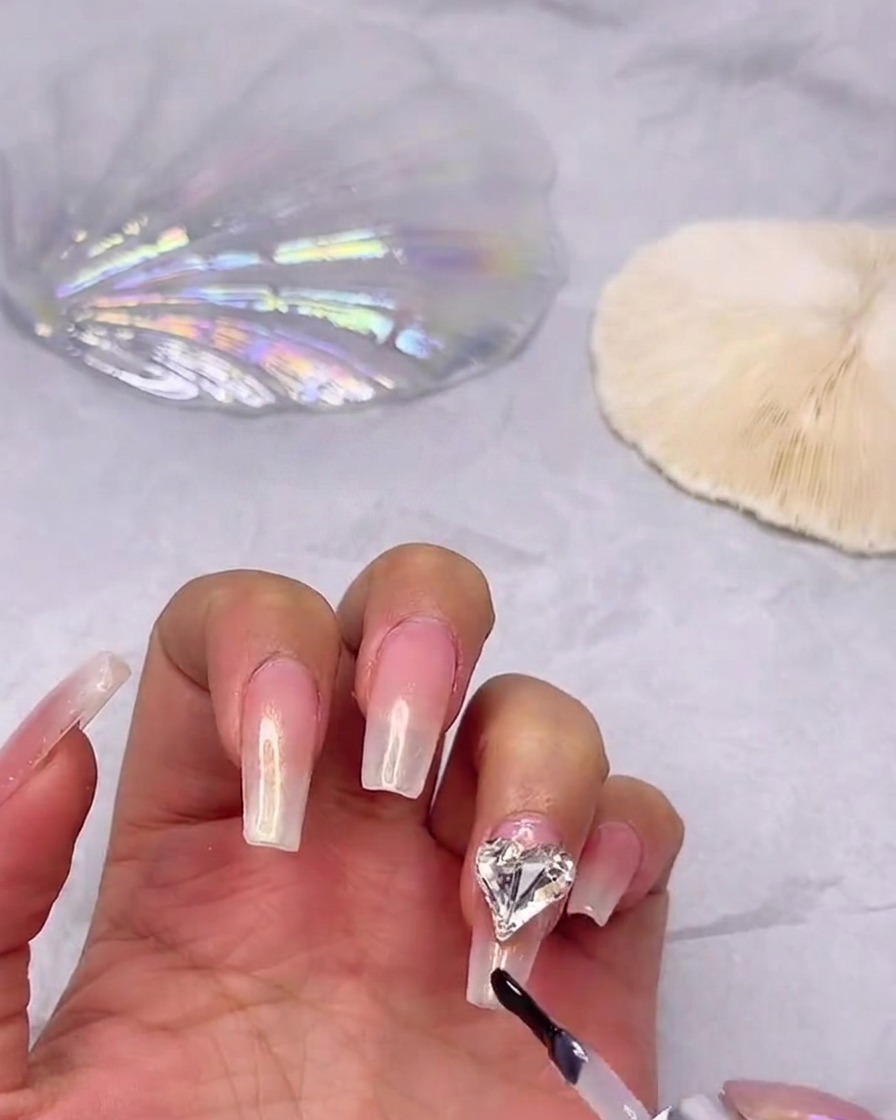 Ein Gel-Pinsel trägt glänzenden Überlack auf einen Ombré-Nagel auf und arbeitet dabei sorgfältig um einen befestigten Herz-Strassstein herum.