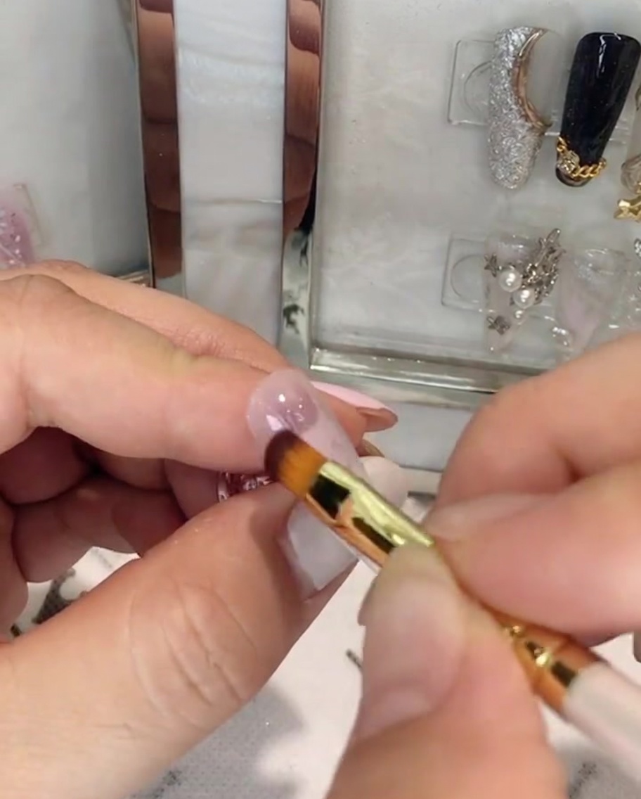 Eine Hand hält einen Nagelpinsel und trägt pinkes Gel auf die obere Hälfte eines mandelförmigen Nagels auf.