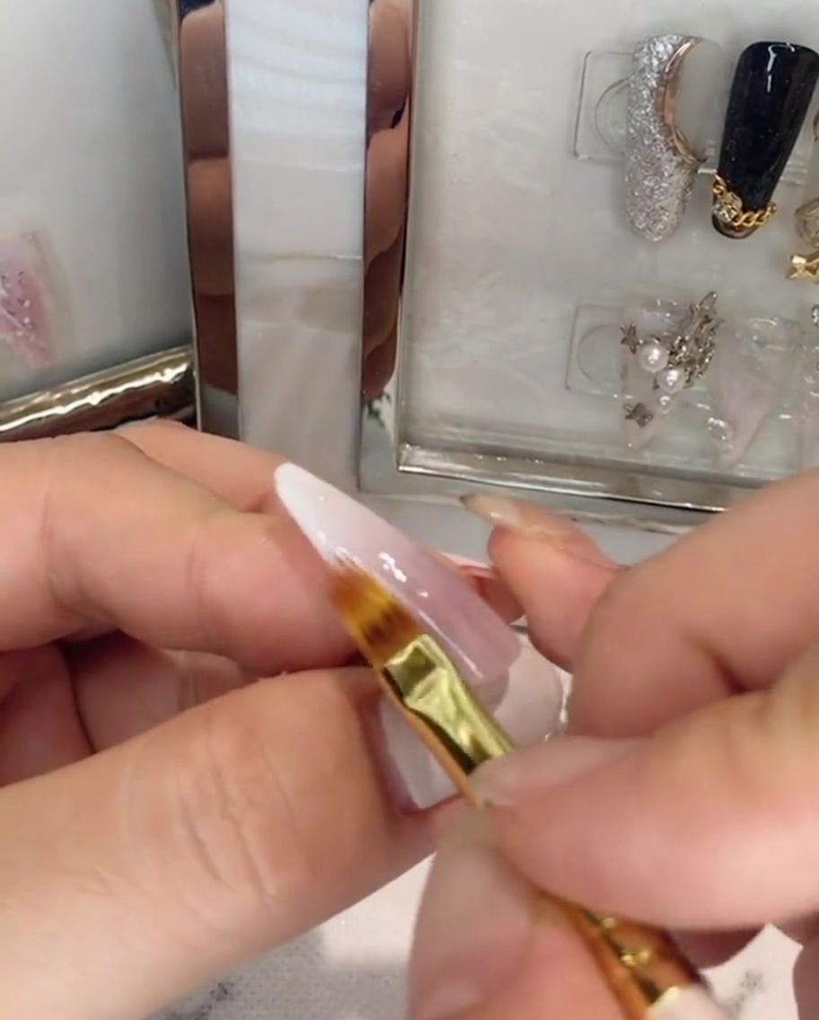 Ein Pinsel trägt einen klaren Topcoat über einen rosa-weißen Ombré-Nagel auf.