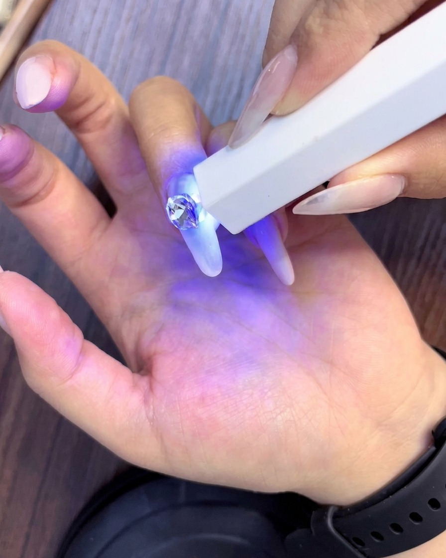 Eine handgeführte UV-Lampe härtet neu platzierte Strasssteine aus, die den Hauptkristall auf einem mandelförmigen Nagel umgeben.