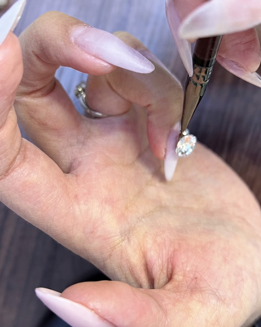 Ein feines Präzisionswerkzeug platziert winzige runde Strasssteine in die kleinen Lücken unter einem großen Nagelkristall.