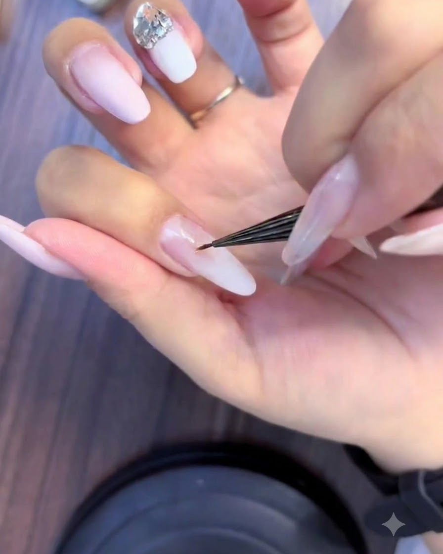 Nahaufnahme einer Nageldesignerin, die mit einem feinen Pinsel kleine Strasssteine zu einem Kristall-Cluster auf einem rosa-weißen Ombré-Nagel anordnet.