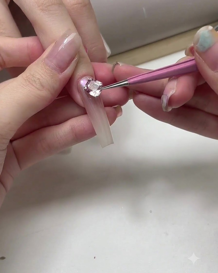 Eine Nahaufnahme einer Nageldesignerin, die mit einem dünnen Pinsel und silbernem Gel filigrane geschwungene Umrisse nahe eines großen Kristalls auf einem Nagel zieht.