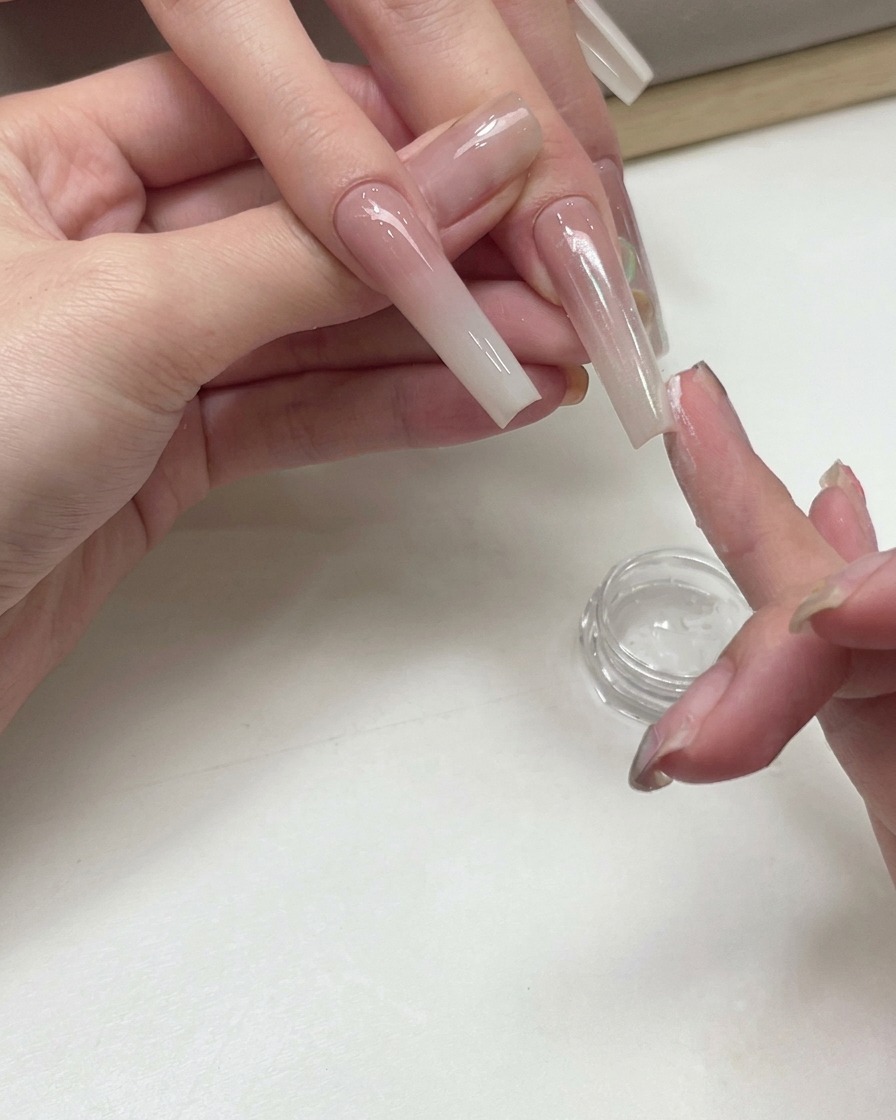 Eine Nageldesignerin verwendet eine Fingerspitze, um schimmerndes Weißpfirsich-Pigmentpuder auf die Oberfläche eines verlängerten Ombré-Nagels zu tupfen.
