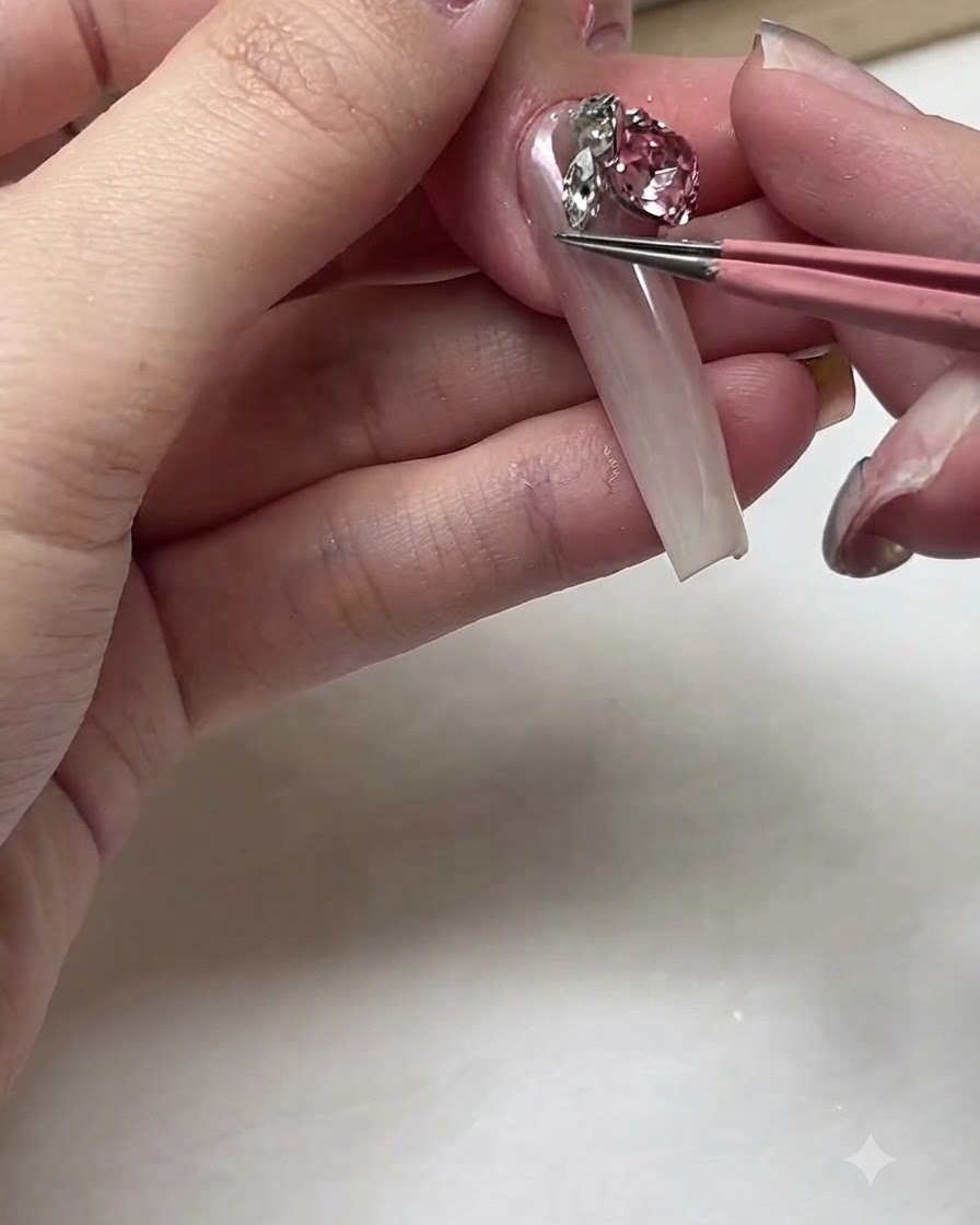 Ein Paar rosa Pinzetten platziert einen großen, funkelnden rosa Kristallstrassstein auf ein langes Nagelbett.