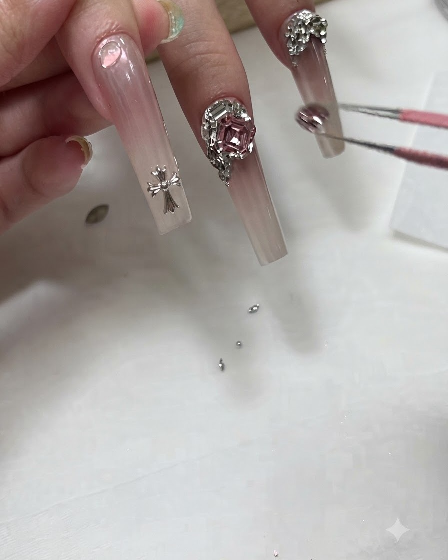 Eine Nageldesignerin verwendet eine Pinzette, um einen kleinen Kristall-Charm nahe der Basis eines langen Ombré-Nagels zu platzieren.