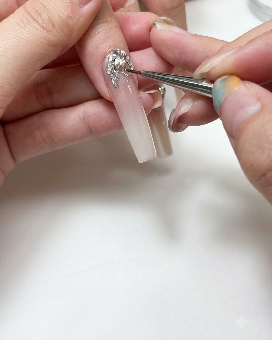 Eine Nahaufnahme einer Nageldesignerin, die mit einem feinen Detailpinsel silberne Gelinien um klare Strasssteine auf einem langen, mit Ombré verlängerten Nagel malt.