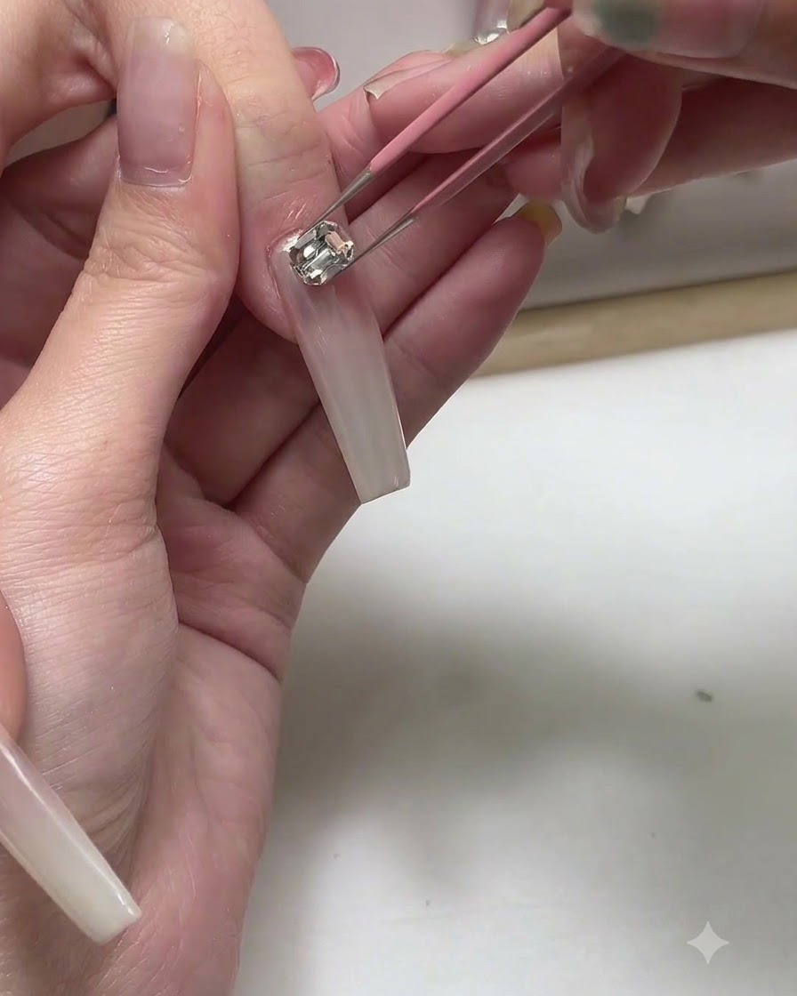 Ein Paar Pinzetten platziert ein kleines Metall-Charm auf einem langen nude-pinken Gel-Nagel.