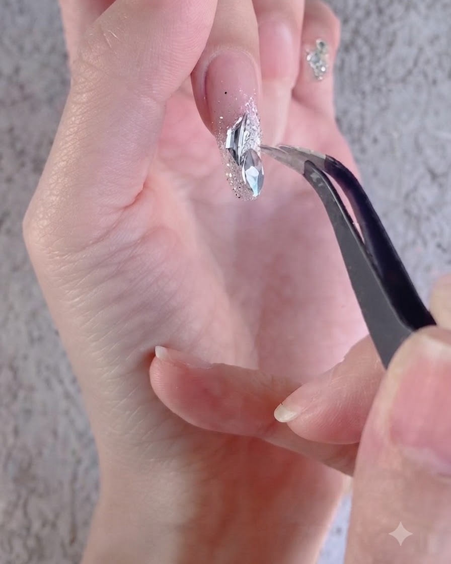 Eine Hand verwendet eine Pinzette, um weisse, unregelmässige Strasssteine auf einem verzierten Nagel anzuordnen.