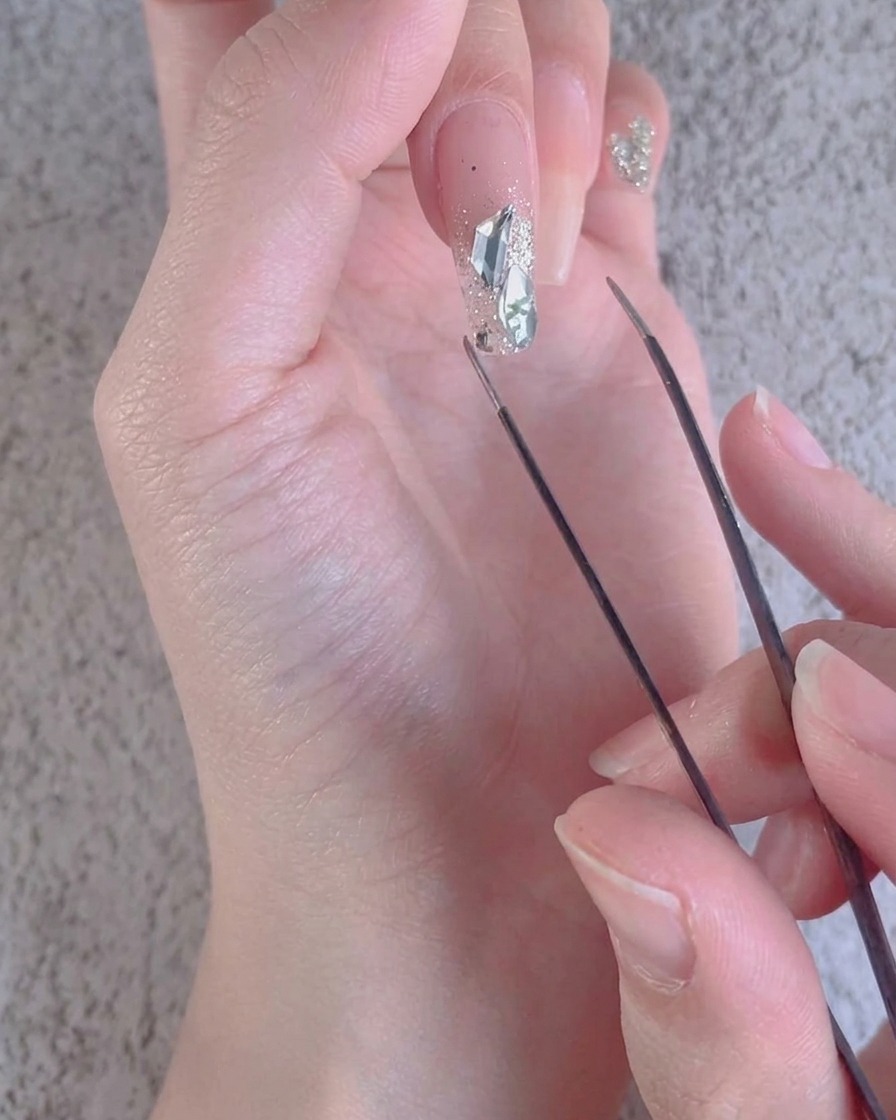 Nahaufnahme einer Pinzette, die kleine zerkleinerte Diamantakzente um grössere Steine auf einem Nagel anpasst.