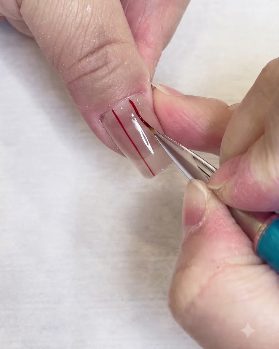 Ein feiner Liner-Pinsel zieht eine zweite parallele rote Vertikallinie auf einem klaren Nagel.