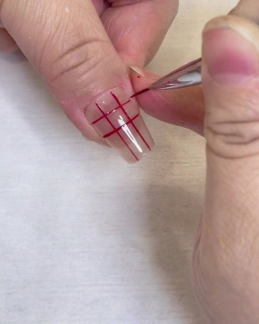 Nahaufnahme eines fertigen roten Gittermusters auf einem klaren Fingernagel, gezeichnet mit einem feinen Liner-Pinsel.
