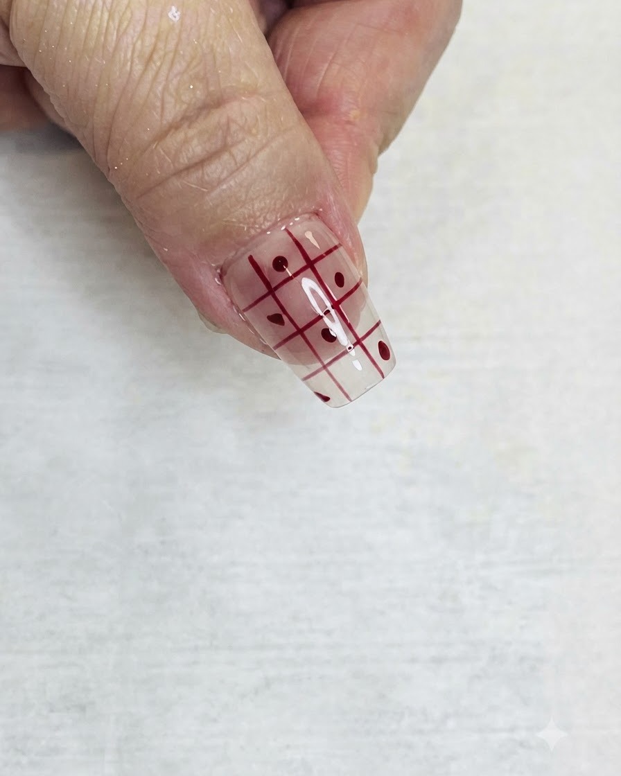 Ein feiner Liner-Pinsel füllt abwechselnde Quadrate eines roten Gittermusters auf einem klaren Nagel mit roter Farbe aus.