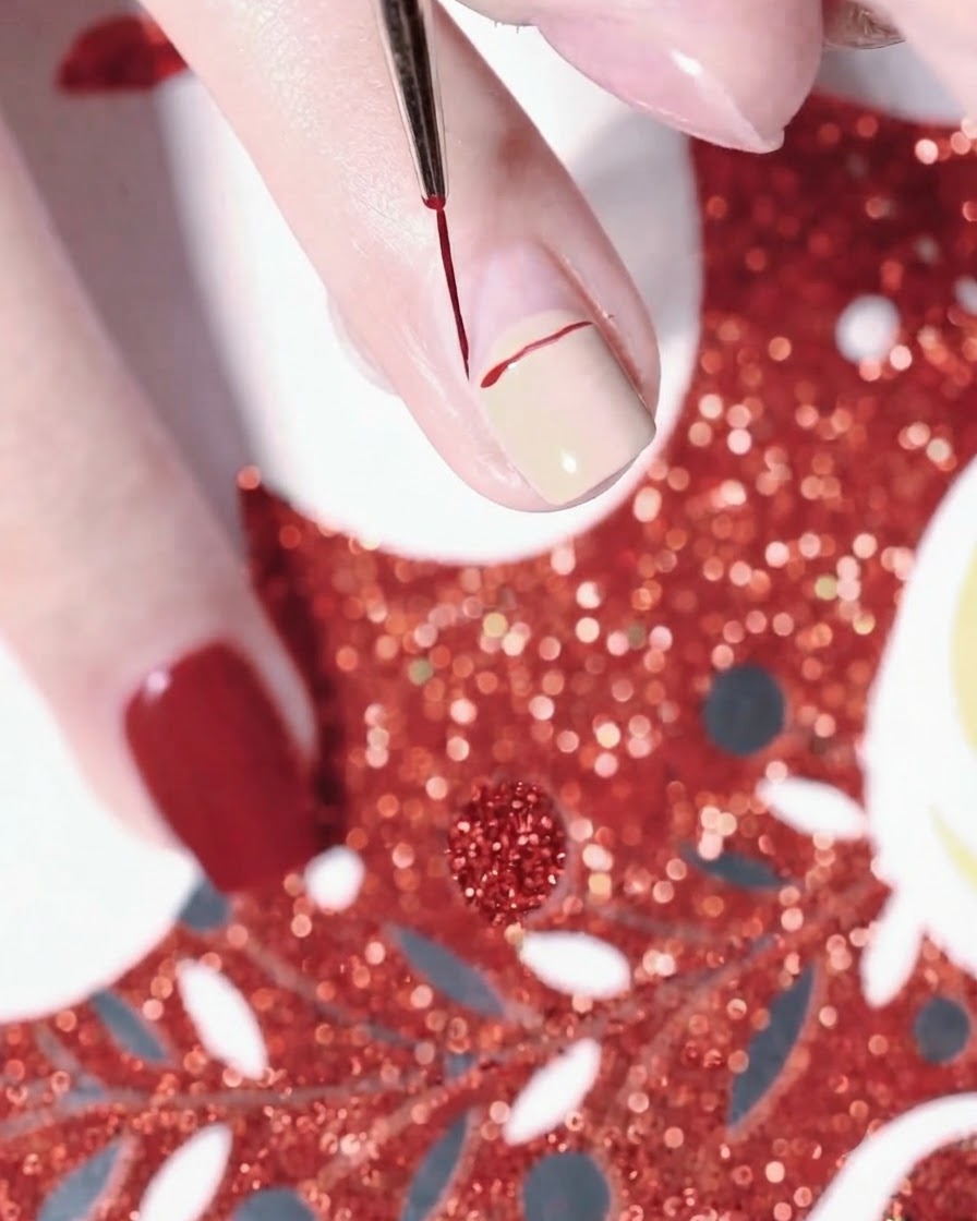 Nahaufnahme einer Hand, die mit einem feinen Liner-Pinsel eine rote, geschwungene Linie für eine Weihnachtsmütze auf einen nude-lackierten Fingernagel zeichnet.