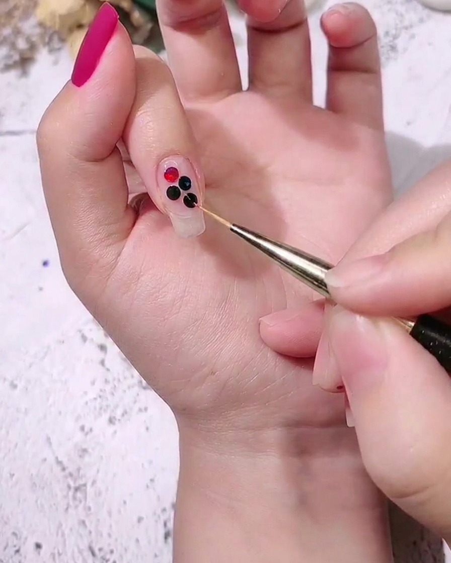 Nahaufnahme einer Hand, die mittelgroße farbige Punkt-Pailletten mit einem Detailpinsel neben größeren auf einen Nagel platziert.