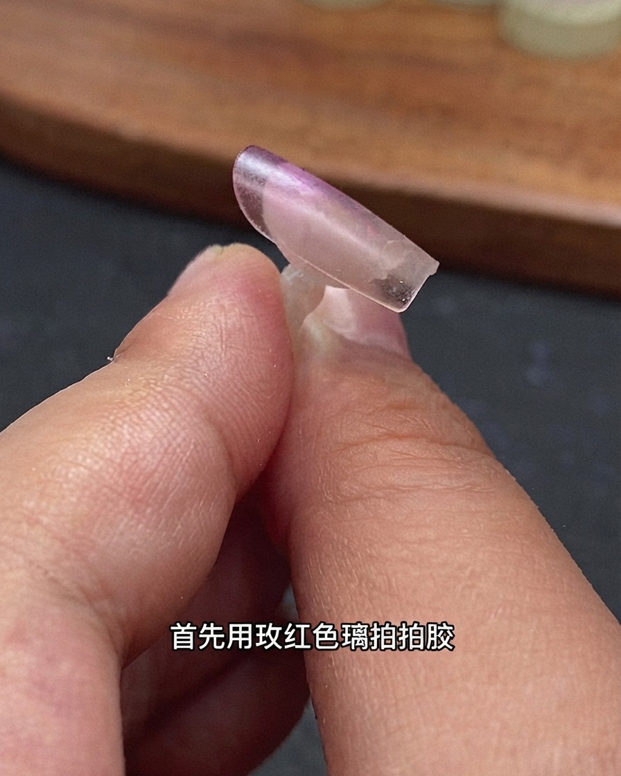 Eine Nahaufnahme einer klaren Nagelverlängerungspitze, die von Fingern gehalten wird; auf die hintere Kante ist rosarotes Glas-Patting-Gel aufgetragen.
