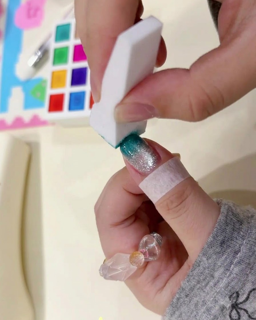Ein weißes Schwämmchen tupft sanft auf die Spitze eines manikürten Nagels, um einen dunkelgrünen Ombré-Effekt aufzubauen.