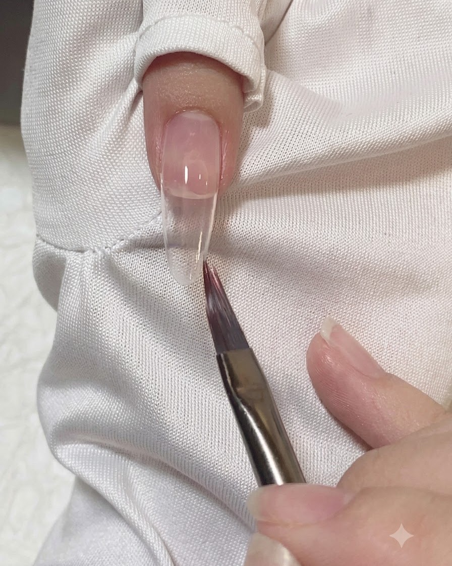 Ein flacher Pinsel glättet und verblendet die Ränder des Rosagels auf einem Nagel.
