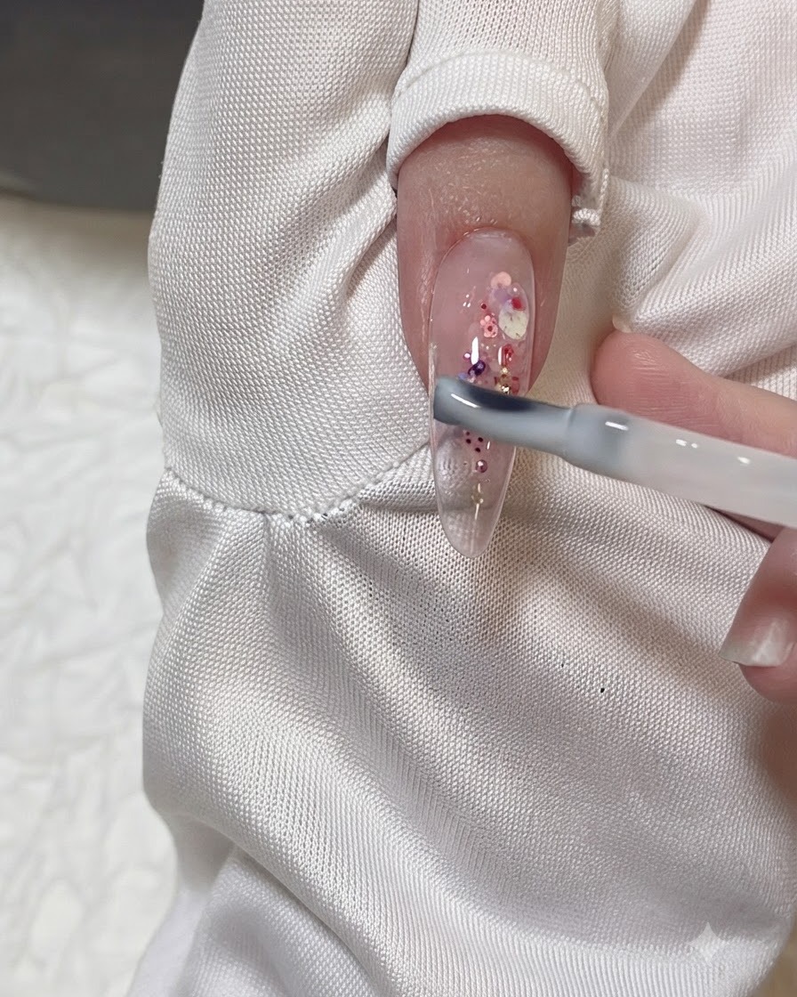 Ein Gelpinsel trägt eine dünne, milchige Gelschicht über einem klaren Mandelnagel auf, der mit rosa und lila Glitzerflocken verziert ist.