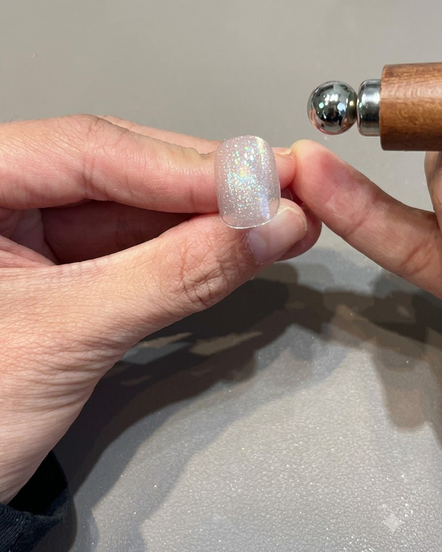 Ein kugelförmiger Magnet wird in die Nähe eines schimmernden, pinkgoldenen Cat-Eye-Gel-Nagels gehalten, um das Lichtband zu formen.