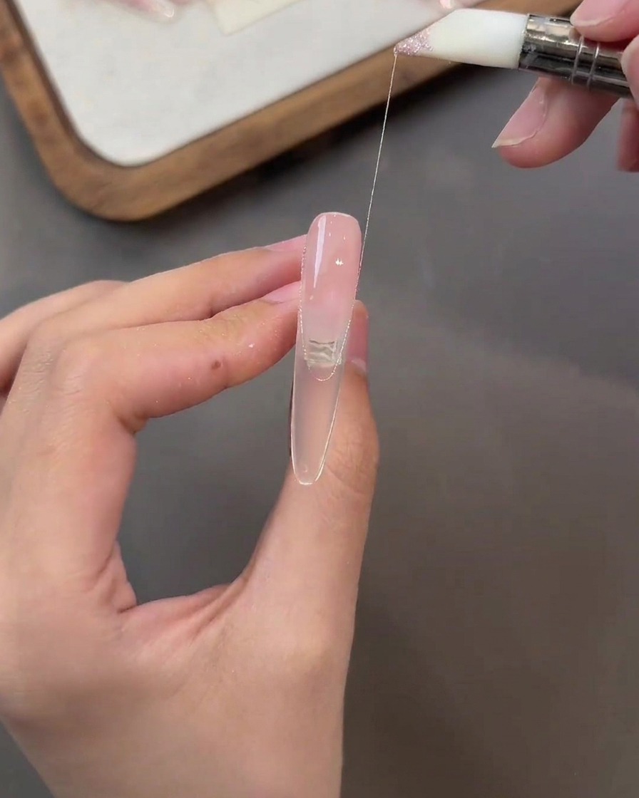 Ein Pinsel zieht einen dünnen Faden aus glitzerndem rosa Gel über die Oberfläche eines nudefarbenen Nagels.