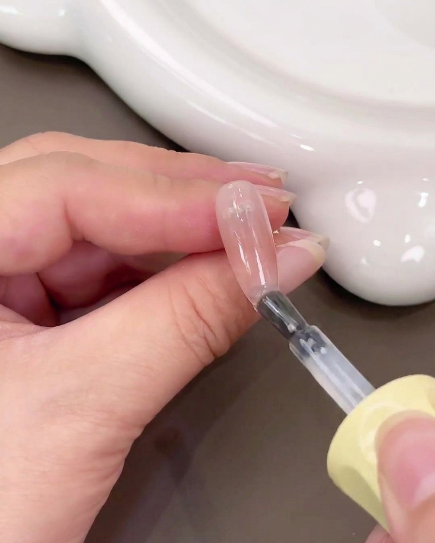 Ein Pinsel trägt klaren Base Gel über einen Nagel mit einem pinkfarbenen Blush-Verlauf auf.