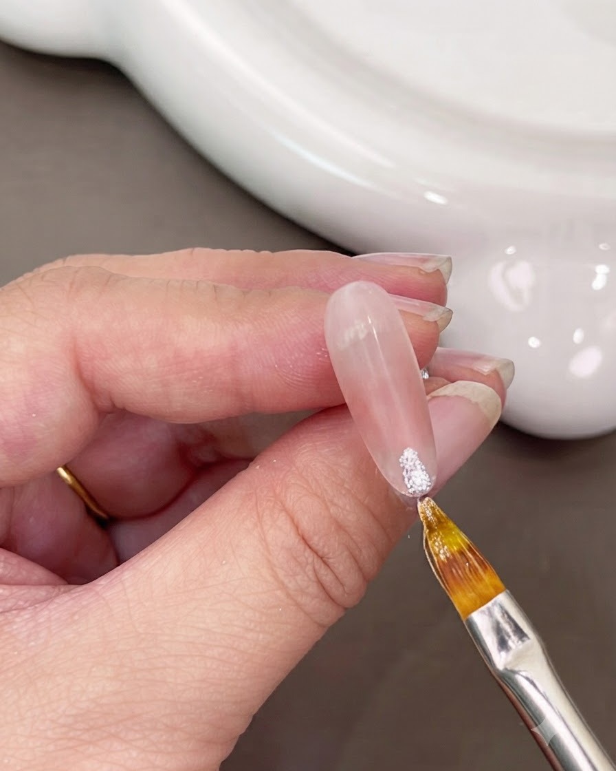 Ein feiner Pinsel trägt glitzerndes weißes Gel auf die Nagelspitze auf.
