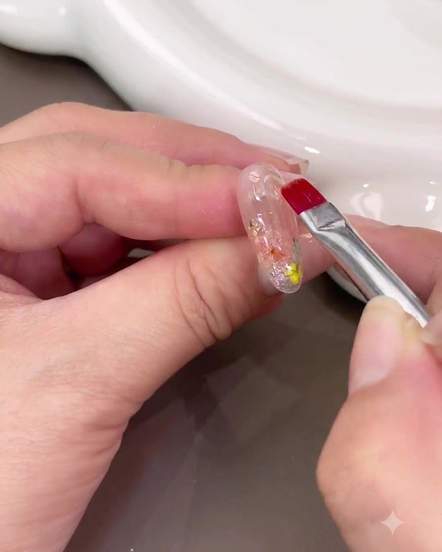 Ein Nagelpinsel trägt eine dicke Schicht klares Builder Gels über das Trockenblumen-Nail-Art auf.