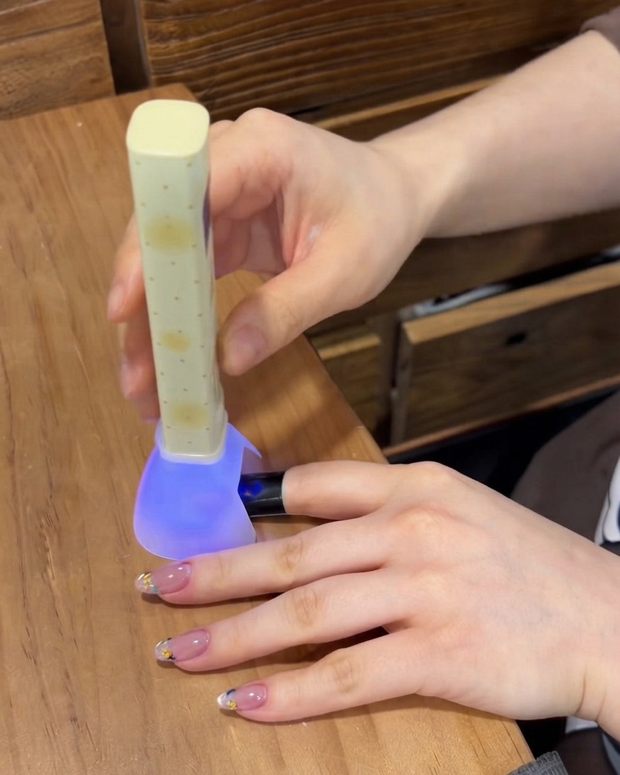 Eine tragbare lila UV/LED-Nagellampe, die das Gel auf einem Fingernagel aushärtet, während andere manikürte Finger auf dem Tisch ruhen.