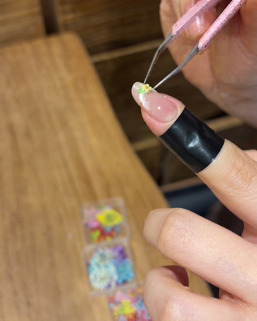 Eine Präzisionspinzette platziert eine winzige gelbe Trockenblume auf der nassen Geloberfläche eines Fingernagels.