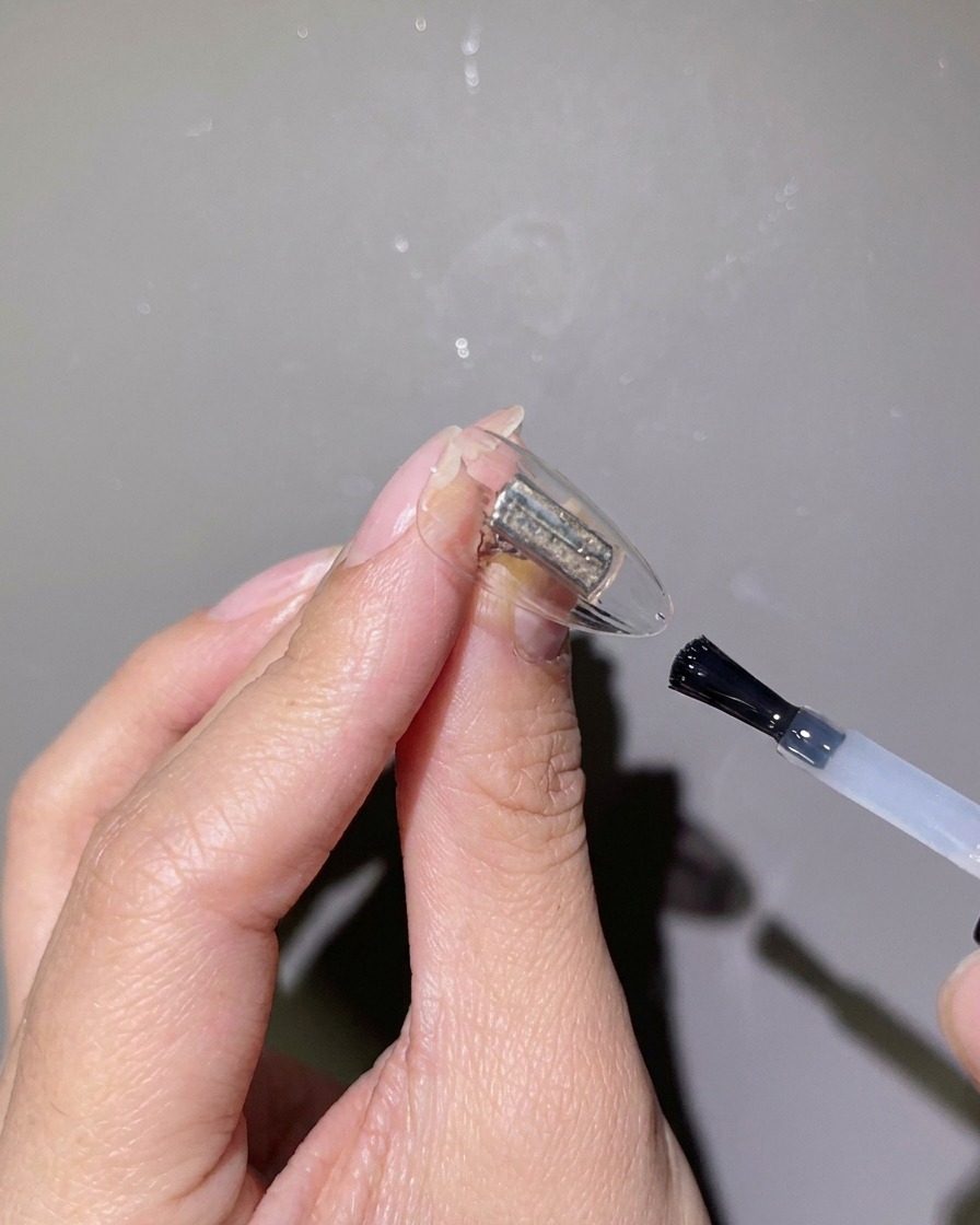 Eine Nahaufnahme eines Pinsel, der klares Verstärkungsgel auf eine klare Nagelspitze aufträgt.