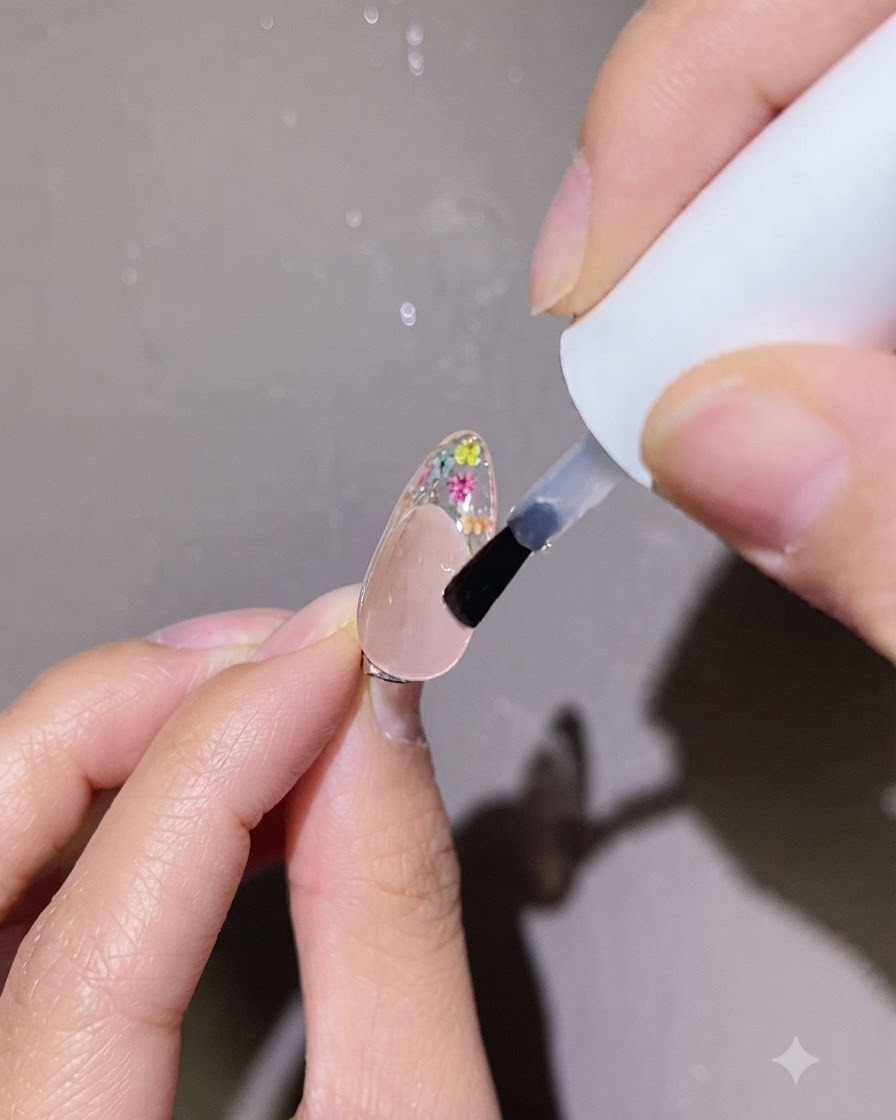 Ein kleiner Pinsel trägt einen präzisen Tropfen klaren Top Coats auf die florale Nagelkunst auf einem Fingernagel auf.