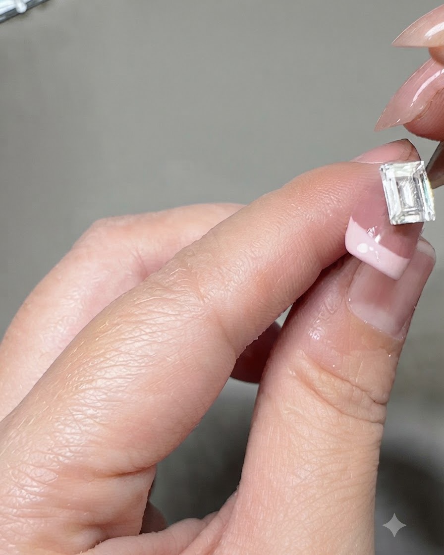 Eine Pinzette setzt einen großen rechteckigen Kristallgemme auf die Mitte eines French-Maniküre-Nagels.