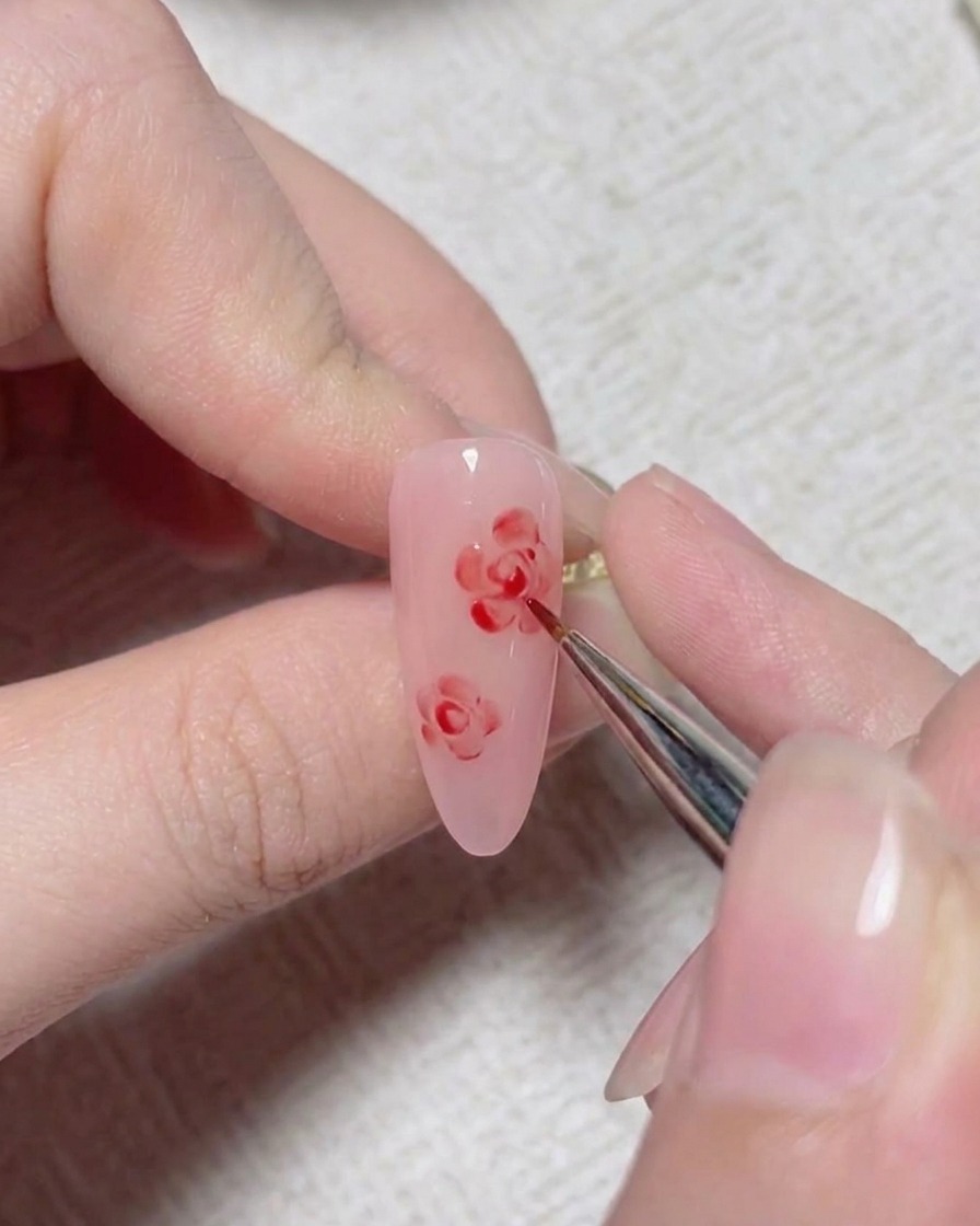 Ein feiner Nailart-Pinsel fügt rote Detaillinien hinzu, um das Zentrum einer gemalten Rose auf einem rosa Nagel zu bilden.