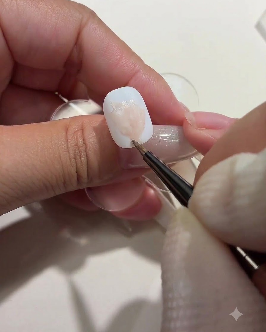 Eine Nail-Artistin trägt mit einem Pinsel weiche rosa Farbflecken auf, die unvollständige Rosenformen auf einem weißen Nagel darstellen.