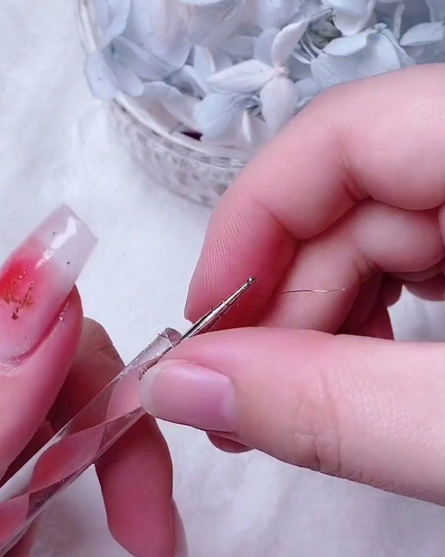 Eine feine Pinzette platziert eine kleine, filigrane Golddraht-Rose in der Mitte eines verzierten Fingernagels.