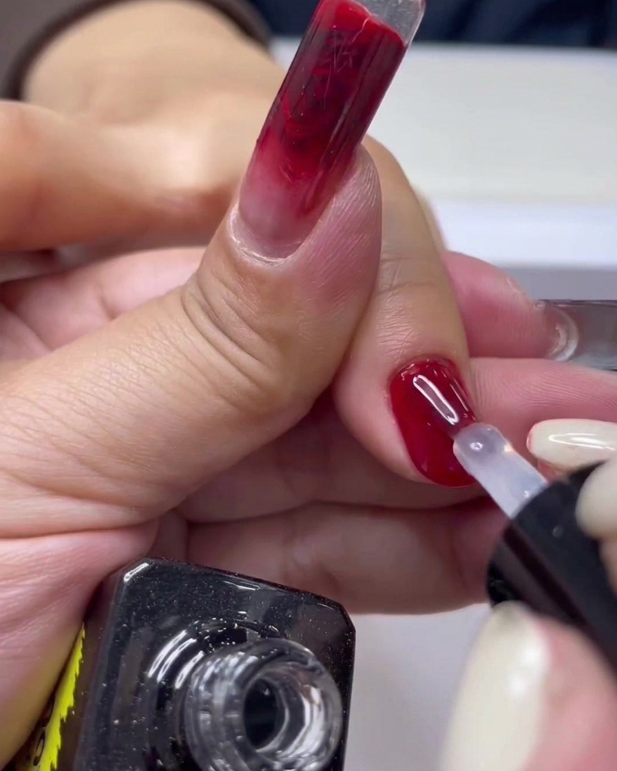 Eine Nahaufnahme eines Nagelpinsels, der klaren Überlack über eine glänzende rote Gel-Maniküre aufträgt.