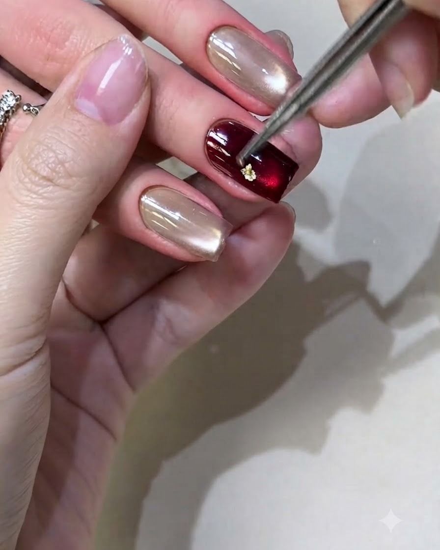 Eine Pinzette platziert kleine Goldfolienstücke auf einem bordeauxfarbenen gel-lackierten Nagel.