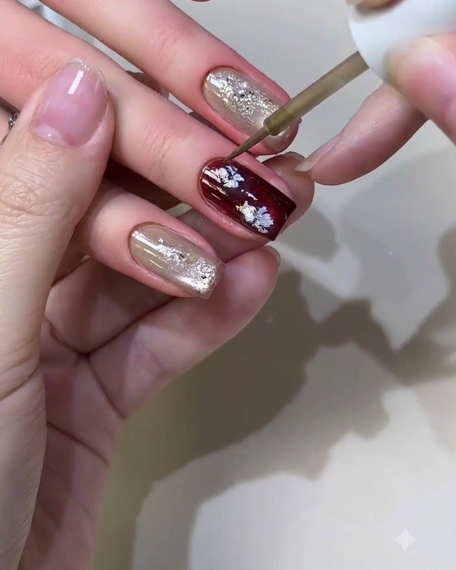 Ein feiner Nail-Art-Pinsel trägt Metallic-Flüssigkeit auf, um kleine, lässige Blumenmuster auf einem bordeauxfarbenen Nagel zu formen.