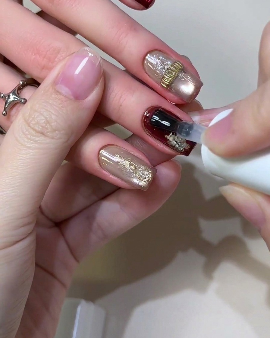 Ein Gelpinsel trägt einen klaren glänzenden Top-Coat-Versiegeler über ein fertiges Retro-Bordeaux- und Blattgold-Nageldesign auf.