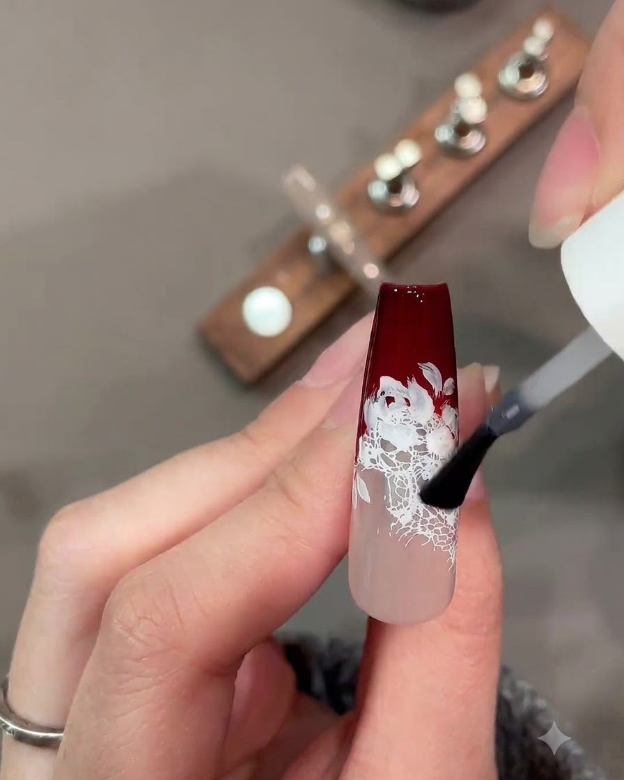 Ein Pinsel trägt einen matten Top-Coat über ein dunkelrotes und weißes Spitzen-Nageldesign auf.