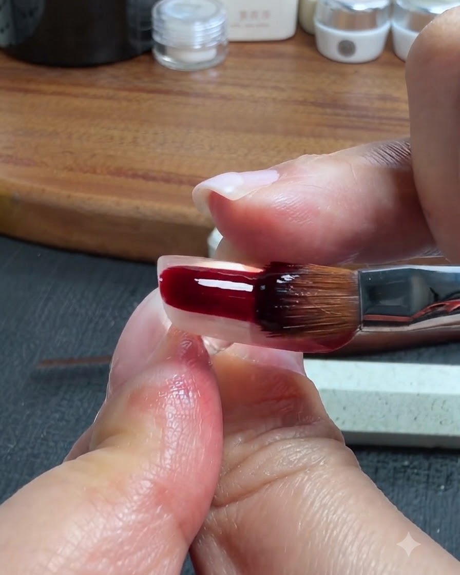 Ein Nagelpinsel trägt ein dunkelrot-braunes, durchsichtiges Gel auf den unteren Teil eines klaren künstlichen Nagels auf.