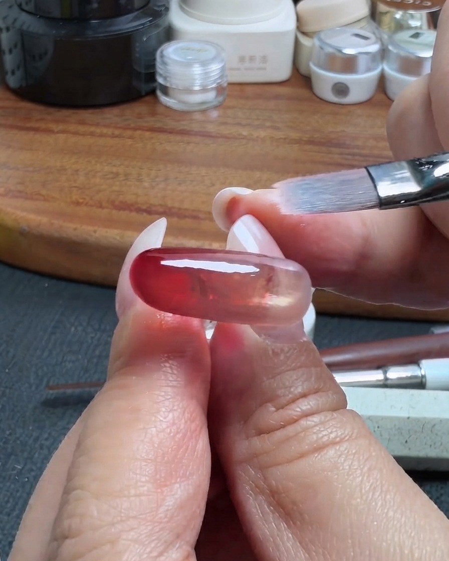 Ein sauberer Make-up-Pinsel verblendet sanft die Kante einer roten, durchsichtigen Gelschicht auf einem Nagel.