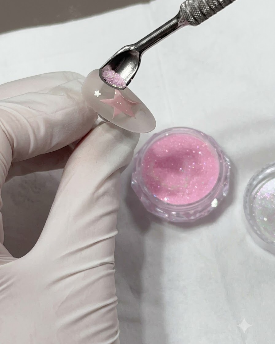 Ein Nagelhautschieber aus Metall hält pinkes Glitzerpuder über einem gemalten Stern auf einem Nagel.